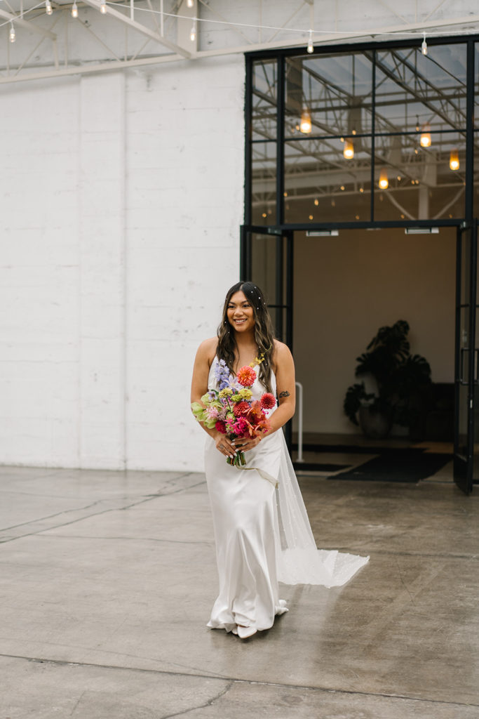 A bride in modern silk dress with colorful bouquet and pearls in hair walks down ceremony aisle at The Revery during cloud themed wedding