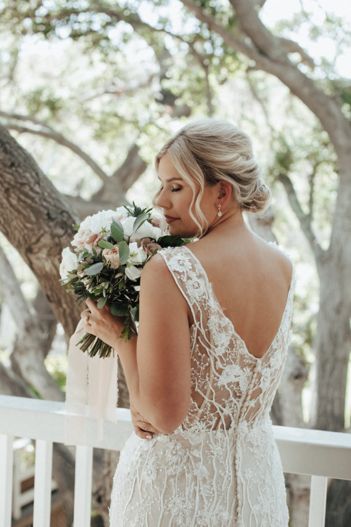 bride in champagne vneck wedding dress with beaded lace overlay standing on balcony of Calamigos Ranch with soft pink and white bridal bouquet