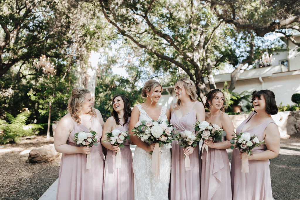 bride in champagne v-neck wedding dress with beaded lace overlay and soft pink and white bridal bouquet stands with bridesmaids in mixed pink dresses for spring wedding at Calamigos Ranch