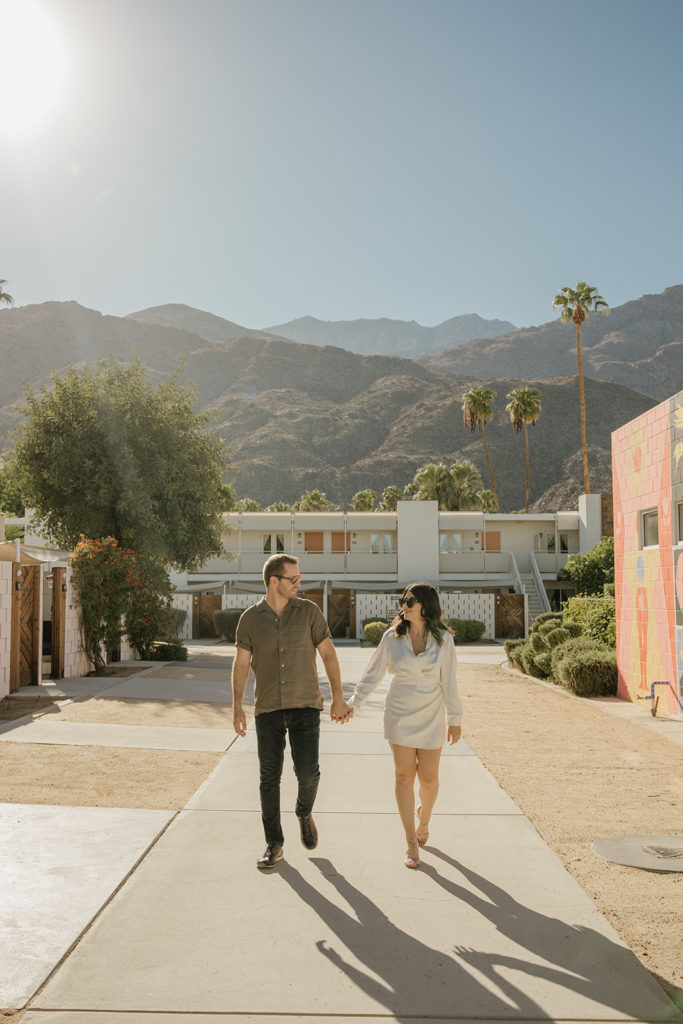 mid-century inspired desert engagement photo session with bride to be in silk mini dress and groom in brown button up walking with mountains in background