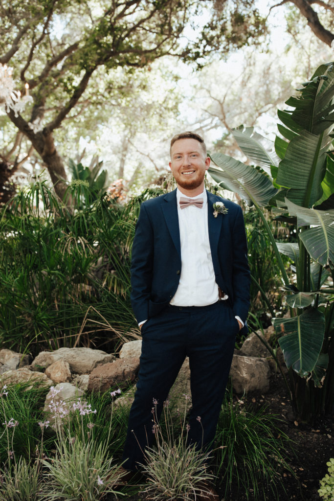 groom in black suit with soft pink bow tie stands in garden