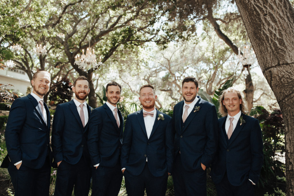groom and groomsmen in black suits with soft pink bow ties stand in garden at Calamigos Ranch