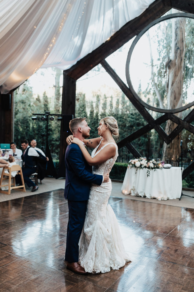 bride in Champagne v-neck dress with beaded lace overlay and groom in dark navy suit with pink bow tie have first dance in The Oak Room at Calamigos Ranch