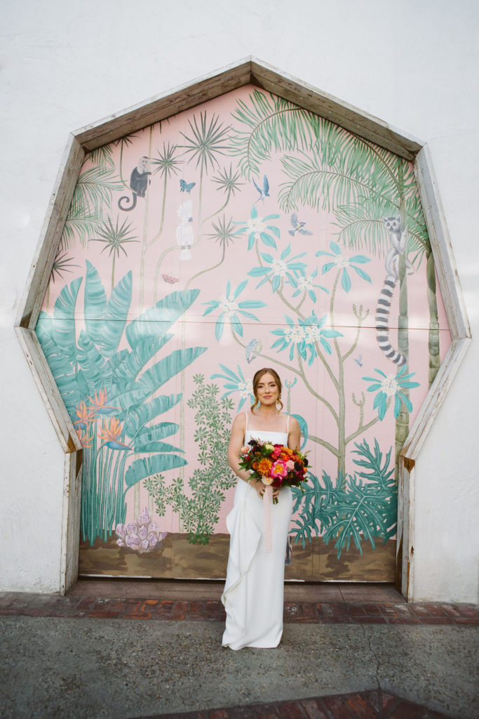 bride in modern square neck spaghetti strap wedding dress holding colorful bouquet in front of pink keyhole wall at Valentine DTLA