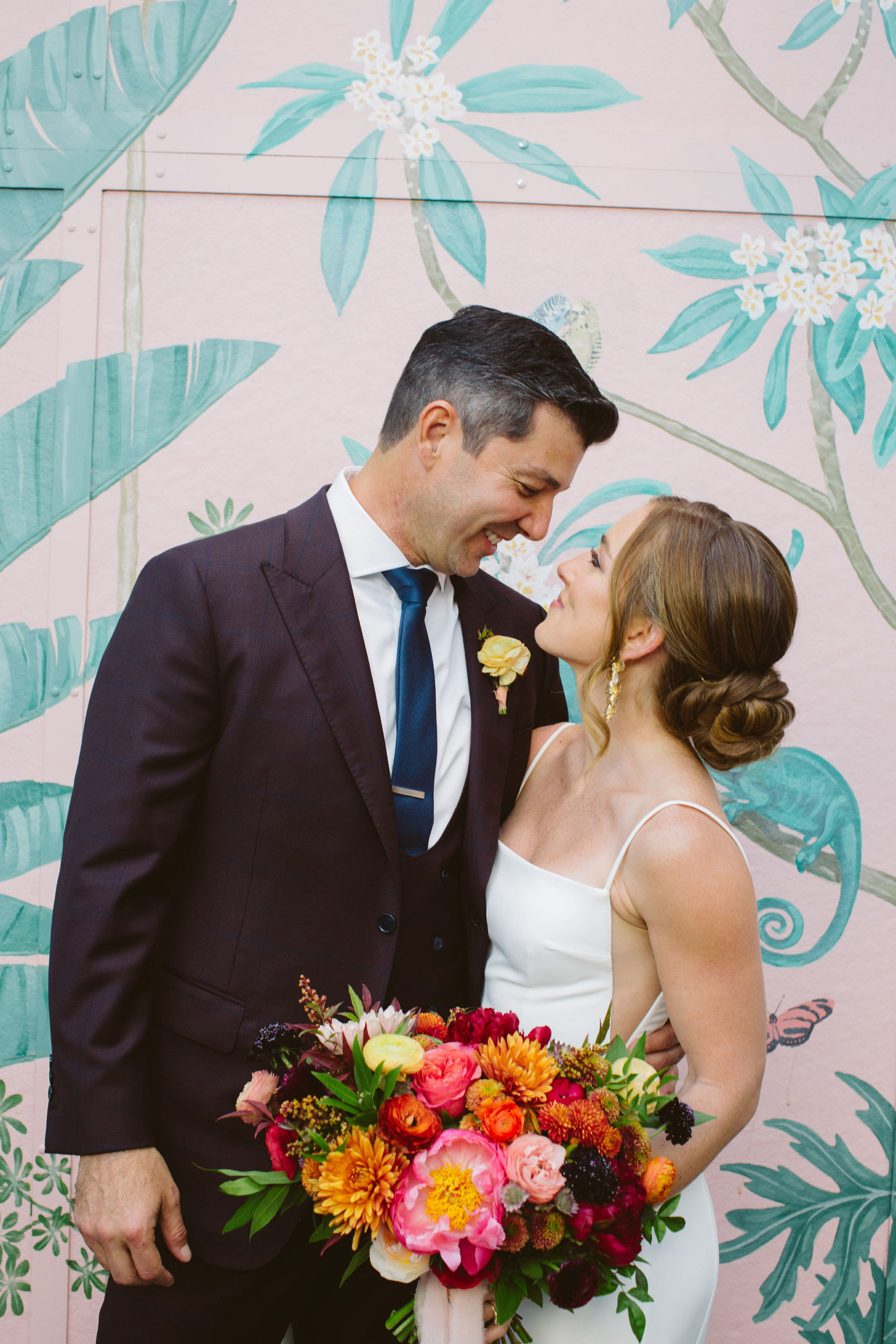 an earth toned wedding with bride in modern spaghetti strap wedding dress and colorful bridal bouquet stands with groom in maroon suit with blue tie in front of pink keyhole wall at the Valentine DTLA 