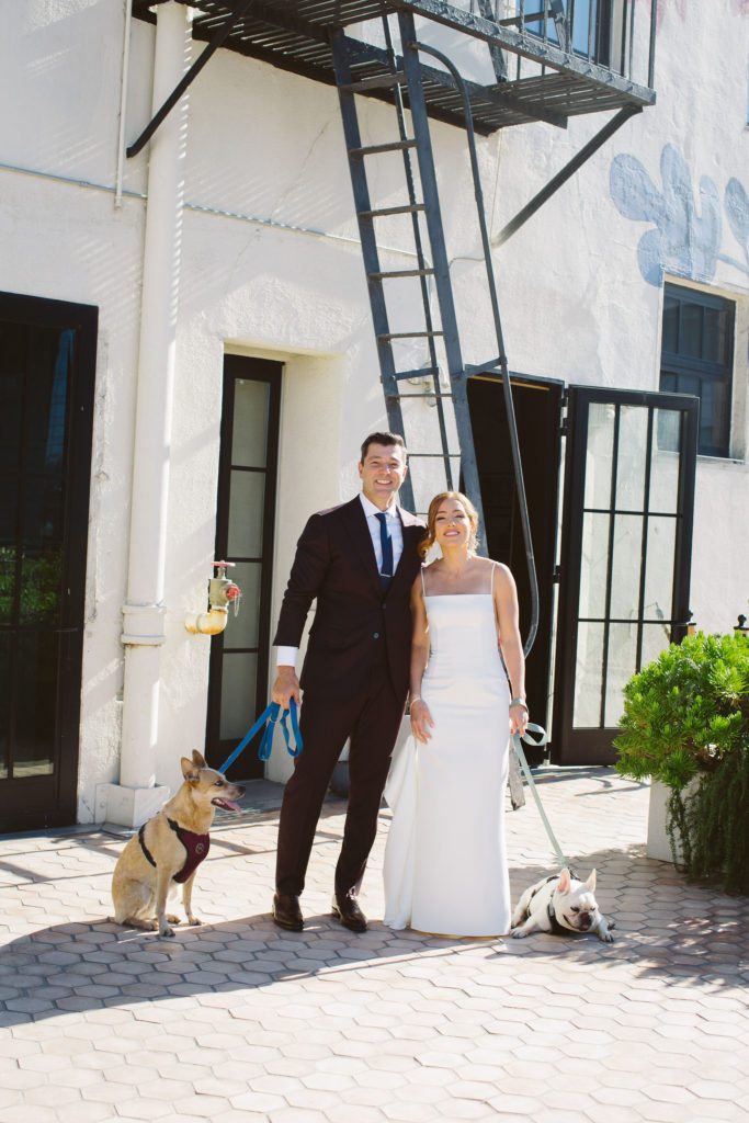 bride in modern square neck spaghetti strap wedding dress and groom in deep plum suit have first look outside at Hotel Figueroa while holding their dogs