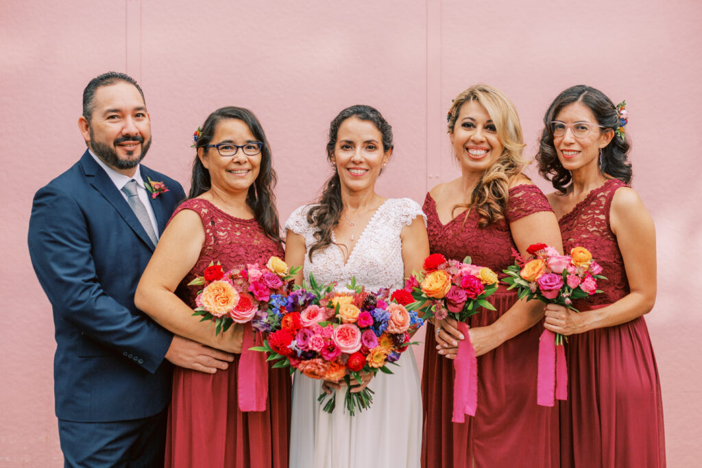 bride in short sleeve lace bodice wedding dress and bright bridal bouquet pose with wedding party in red and blue attire at Grass Room 