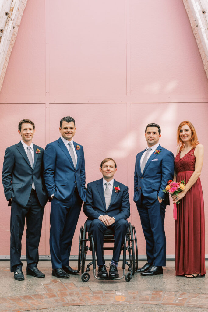 groom in blue suit and colorful boutonniere poses with wedding party in red and blue attire