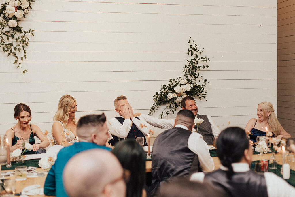 bride and groom laugh during modern wedding reception at Prospect House with wooden oak chairs and tables, dark green linens, beige taper candles and small bud vases