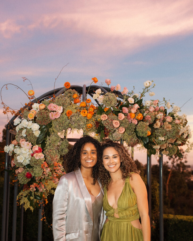 Bride to be in satin green retro inspired dress stands with bride to be in silver satin suit during their sunset themed engagement party in Bel-Air